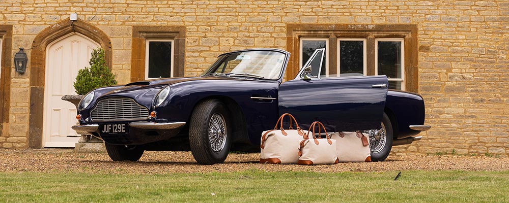 The Tusting Como Collection of Explorer Luggage, set next to a beautify, dark blue Aston Martin car